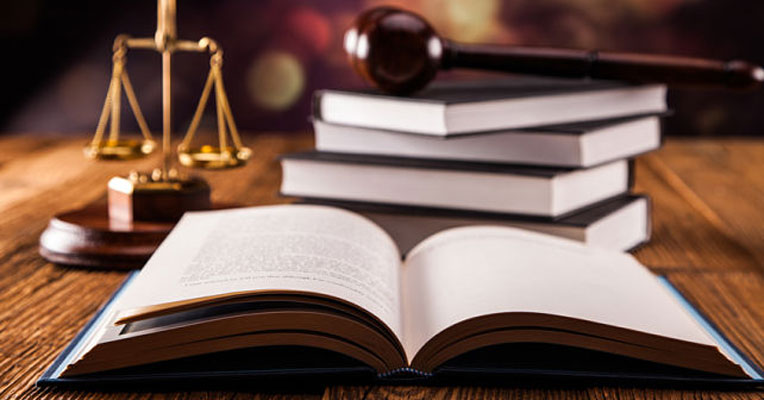 Scales of justice, law books and a judge's gavel stacked on a desk symbolizing legal defense.